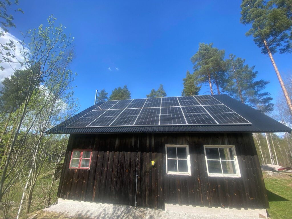 Ett litet trähus med svärtade väggar i Grycksbo har solceller installerat på taket. Omgiven av träd och mot en klarblå himmel en solig dag, är scenen både pittoresk och hållbar.