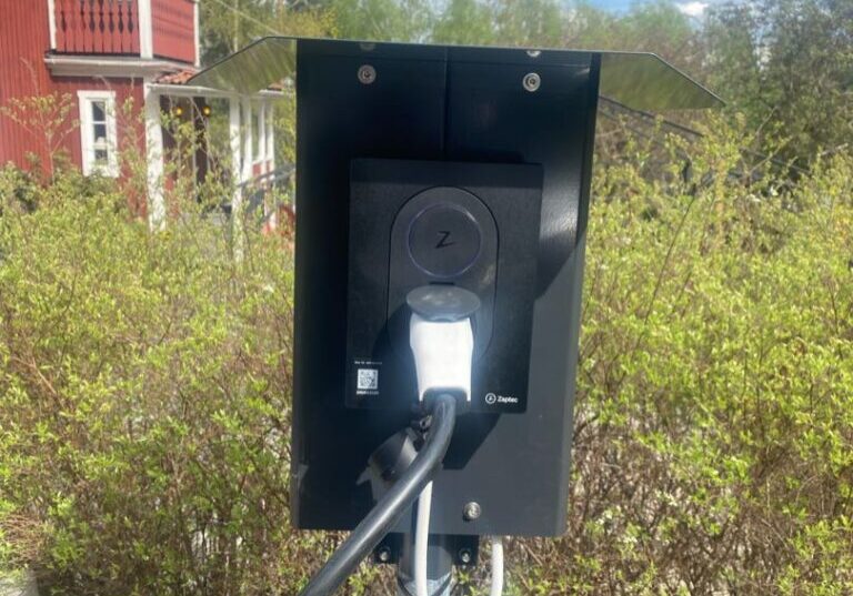 En Laddbox EV-laddstation är installerad utomhus, monterad på en metallstolpe med en laddkabel inkopplad. I bakgrunden finns ett rött hus med vit klädsel på Pilbovägen i Falun, omgivet av grönska och under en klar, blå himmel med spridda moln.