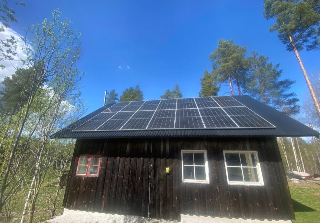 Ett litet trähus med svärtade väggar i Grycksbo har solceller installerat på taket. Omgiven av träd och mot en klarblå himmel en solig dag, är scenen både pittoresk och hållbar.
